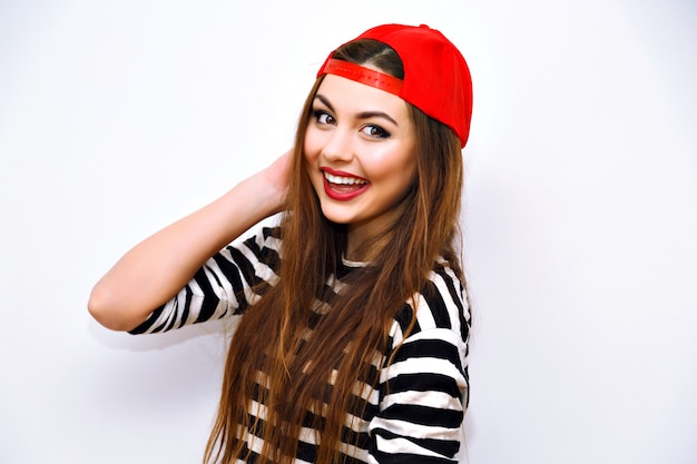 Foto gratuita close up ritratto di stile di vita di moda al coperto di fresca bella giovane donna bruna, capelli lunghi incredibili, trucco luminoso, cappello rosso e maglietta a righe, grande sorriso incredibile, viso carino, immagine urbana con flash.