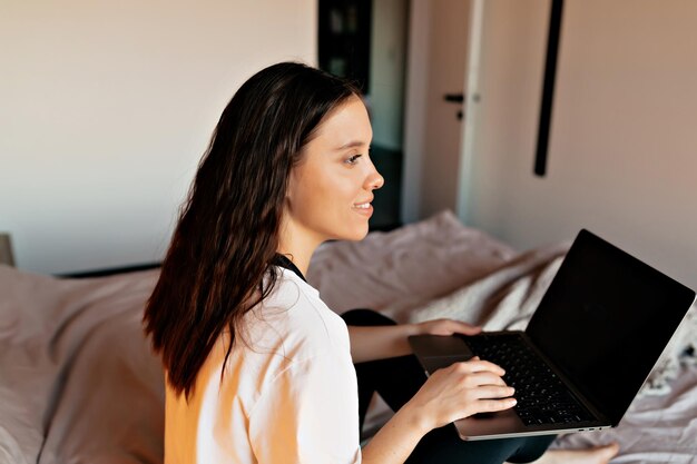 흰 셔츠를 입은 검은 머리 소녀의 실내 사진을 닫고 집에서 침대에 노트북을 들고 일하고 있다