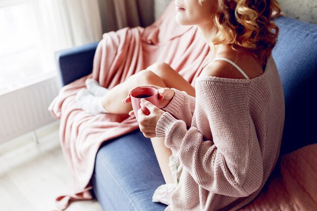 Close up indoor portrait of graceful blonde woman enjoying smell of cappuccino  , dreaming and looking into the window.  Wearing pink knitted sweater.