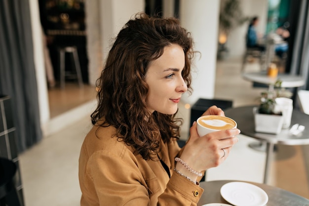 Foto gratuita primo piano ritratto al coperto di una donna piuttosto affascinante con i capelli ricci che indossa una camicia marrone tiene in mano una tazza di caffè e guarda avanti ricreazione colazione mattutina