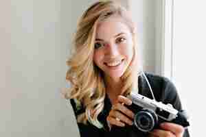 Free photo close up indoor portrait of smiling charming girl with blonde curly hair posing with retro camera while sitting on the window