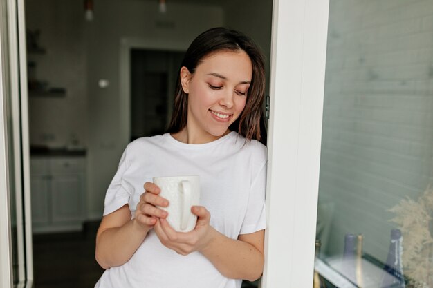Close up indoor portrait of smiling european woman with dark hair wearing white t-shirt drinking coffee in the morning at the kitchen.