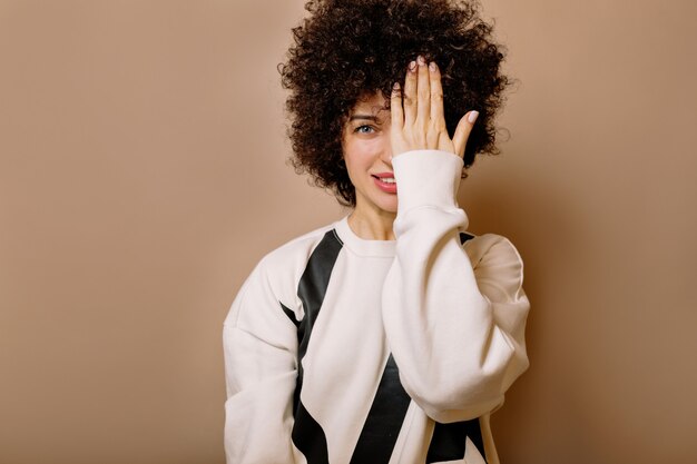 Close up inside portrait of charming lovable girl with afro hairstyle looking at front with smile and covering face with one hand on beige wall