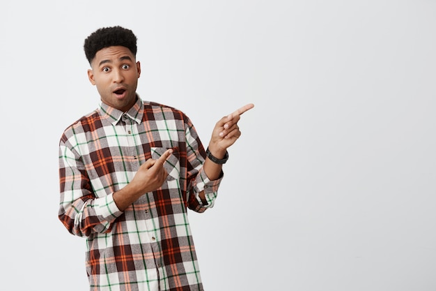 Free photo close up isolated portrait of beautiful young dark-skinned african student guy with fashionable shirt pointing aside on white wall with opened mouth and surprised expression