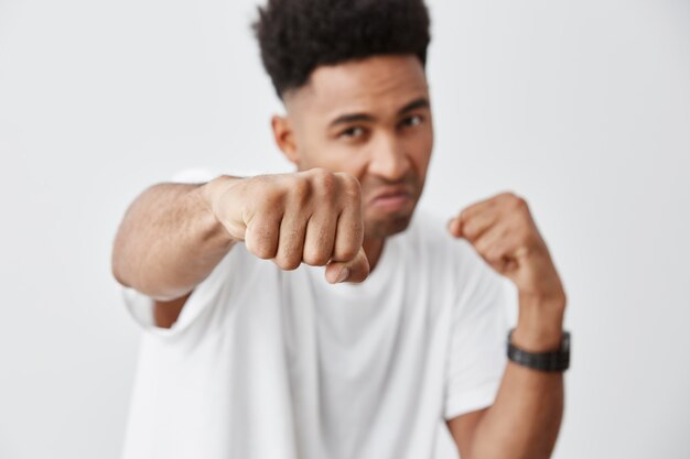 Close up isolated portrait of young attractive black-skinned man with afro haircut in white casual t shirt posing with aggressive face expression, showing fist in camera