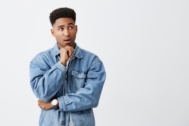 Free photo close up isolated portrait of young beautiful dark-skinned man with afro hairstyle in denim jacket touching chin with hand, looking aside with thoughtful expression. copy space
