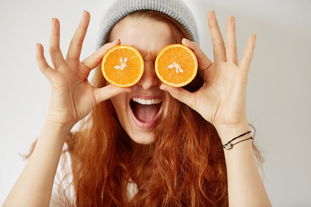 Close up isolated portrait of young redhead woman holding halved oranges at her eyes