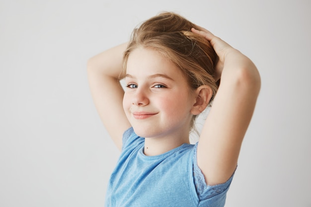 Free photo close up of joyful little girl in blue t-shirt looking , brightfully smiling and holding blonde long hair with hands. copy space.