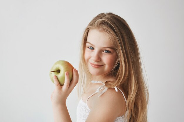 Close up of joyful small light-haired girl with blue eyes in white dress holding apple in hand, smiling brightfully .