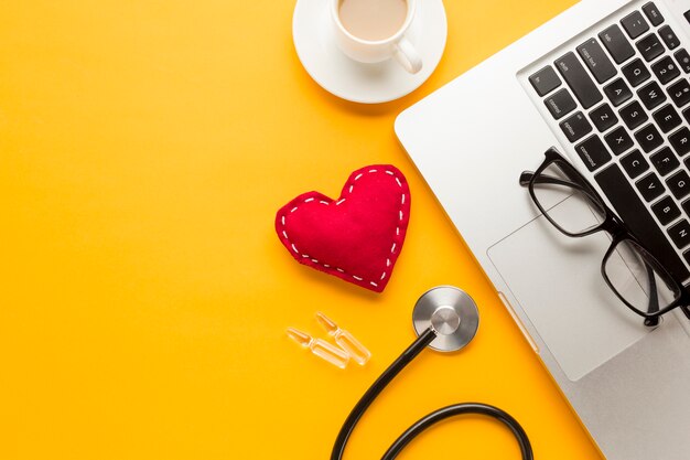 Close-up of laptop keyboard; stitched cloth toy; coffee cup; ampoule; stethoscope against yellow desk