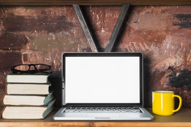 Close-up of laptop; stacked books; spectacles and cup on wooden shelf