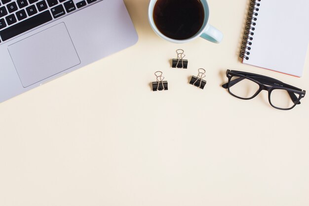 Close-up of laptop; tea cup; paperclip; eyeglasses and spiral notepad on beige backdrop