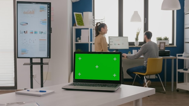 Close up of laptop with green screen at empty desk in office. Computer with chroma key on display, mock up template and isolated background. Device with chroma-key copy space app.