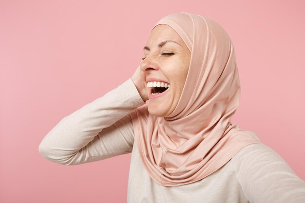 Close up laughing funny young arabian muslim woman in hijab light clothes posing isolated on pink background. people religious lifestyle concept. mock up copy space. doing selfie shot on mobile phone.