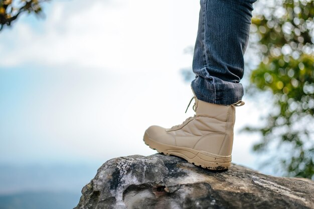 Close up Leg of hiking man in jeans with trekking sneakers placed on rocky mountain peak in the natural forest blue sky on background copy space