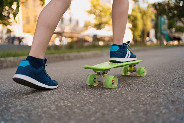 モーションで緑のスケートボードに乗って青いスニーカーで足を閉じます。若者のアクティブな都会のライフスタイル、トレーニング、趣味、活動。子供のためのアクティブなアウトドアスポーツ。子供のスケートボード。