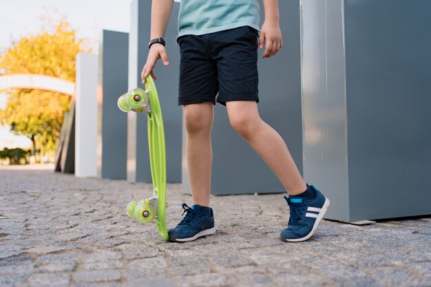 Close up legs in blue sneakers with green skateboard. Active urban lifestyle of youth, training, hobby, activity. Active outdoor sport for kids. Child skateboarding.