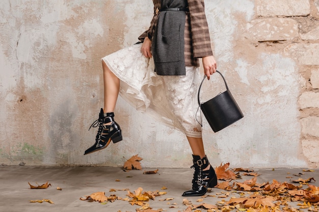 Close up legs in boots of stylish woman in jacket walking against wall in street