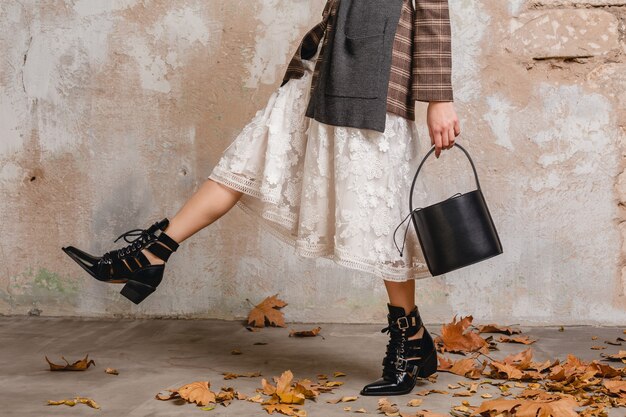 Close up legs in boots of stylish woman in jacket walking against wall in street