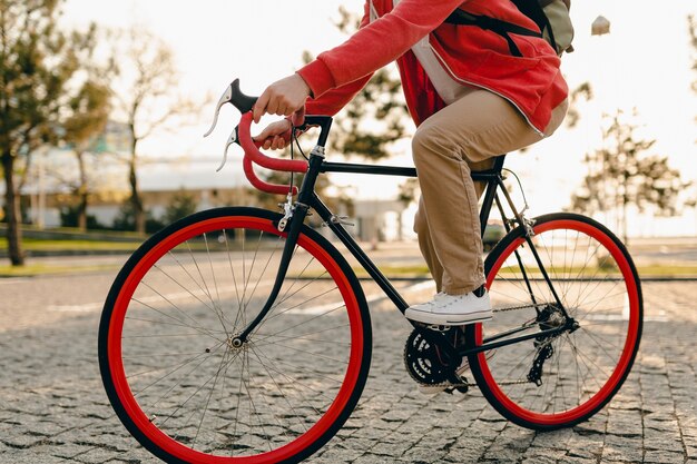 Close up legs in sneakers and hands on steering wheel of hipster style bearded man in red hoodie and beige trousers riding alone with backpack on bicycle healthy active lifestyle traveler backpacker