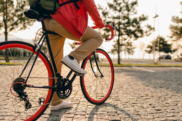 スニーカーで足を閉じ、赤いパーカーとベージュのズボンでヒップスタースタイルのひげを生やした男のハンドルに手を自転車でバックパックと一緒に乗る健康的なアクティブライフスタイルトラベラーバックパッカー