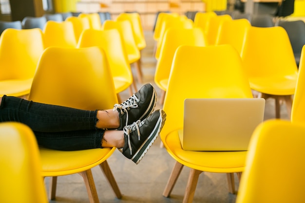 Закройте ноги молодой стильной женщины, сидящей в лекционном зале с ноутбуком, в классе с множеством желтых стульев, кроссовками, модной тенденцией обуви
