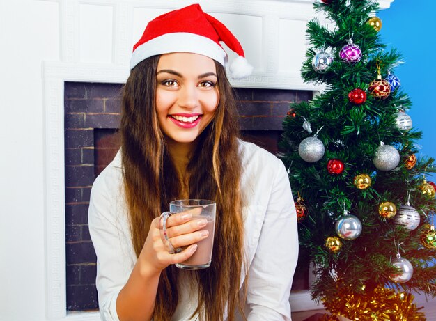 Close up lifestyle portrait of pretty brunette woman drinking hot chocolate in New year eve, wearing santa hat and sitting near fireplace and decorated Christmas tree. Cozy home atmosphere.
