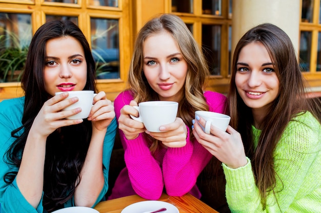 Закройте вверх по портрету образа жизни трех красивых молодых женщин, сидящих в кафе и наслаждающихся горячим тройником. Ношение яркого неонового желтого, розового и синего стильного свитера. Праздники, еда и концепция туризма.