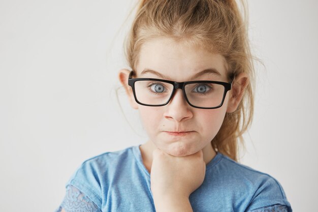 Close up of little cute girl with big blue eyes and light hair looking  with angry face expression and raised eyebrows in glasses.