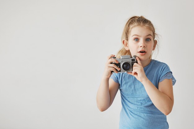 カメラを手で押し、驚いた表情で青いtシャツに光の髪のかわいい女の子のクローズアップ。