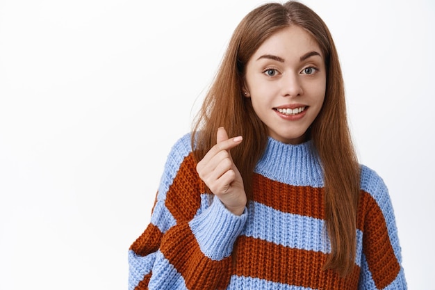 Foto gratuita primo piano di una ragazza adorabile che sorride e mostra il segno del cuore con il dito, ti amo gesto, come qualcosa, in piedi in un maglione su sfondo bianco