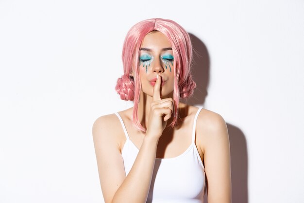 Close-up of lovely girl with pink wig and bright makeup, close eyes and shushing, have surprise or secret, standing.