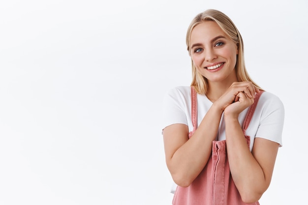 Close-up lovely, tender feminine young woman in pink trendy overalls, t-shirt with tattoos on arms, clench hands together in tender and coquettish pose, smiling happily camera, white background