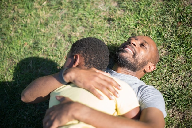 彼の息子を抱き締める愛情のあるアフリカの父のクローズアップ。芝生の地面に横たわっている笑顔の男と夏に彼の胸に巻き毛の少年。親は愛し、世話をし、保護し、一緒に時間を過ごすという概念