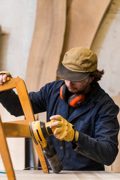 ワークベンチで電動工具で家具を研磨している男性の大工のクローズアップ