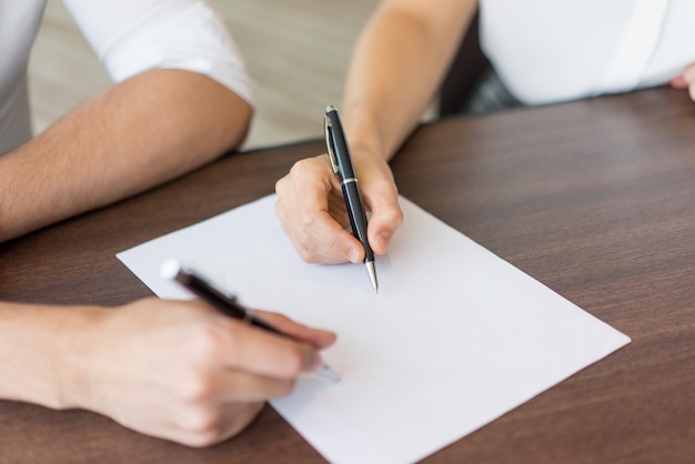 Close-up of male and female hands writing on paper sheet at table. 