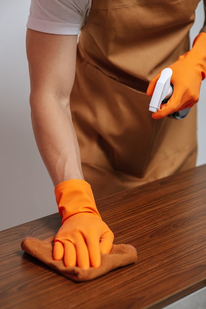 Бесплатное фото Закройте мужскую руку, используя салфетки из ткани на деревянном шкафу с ящиком, и держа бутылку с распылителем, чтобы мыть мебель дома