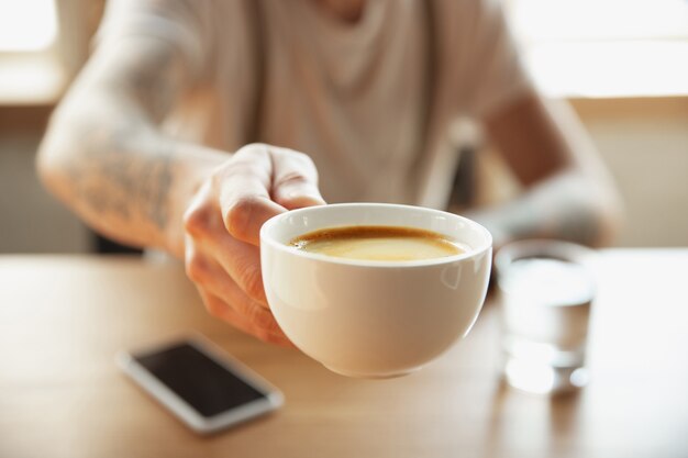 Close up of male hands proposing cup of coffee, sitting at the table with smartphone. Surfing, online shopping, working. Education, freelance, art and business concept. Drinking. Hot aroma drink.