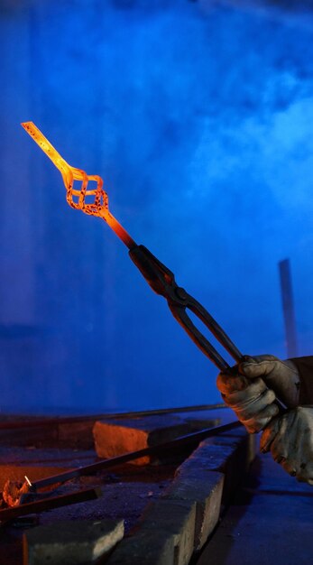 Close up of male hands in safety gloves holding forceps with heated shaped steel Professional blacksmith working with molten metal at forge Concept of handwork and industry