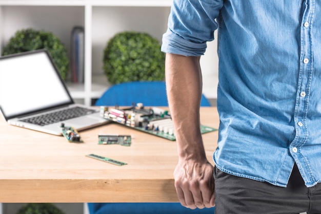 Primo piano del tecnico maschio che si appoggia al bordo della tabella con attrezzature del computer portatile e dell'hardware