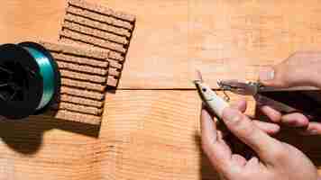 Free photo close-up of a man fixing the hook with plier near the reel and cork board on wooden desk