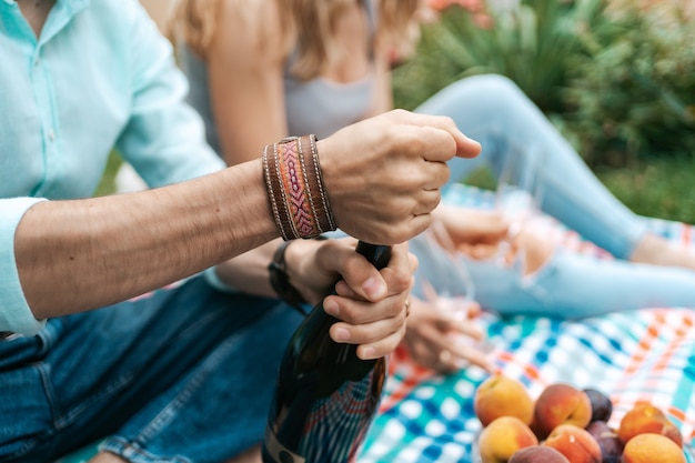 그의 아내가 서로를 즐기고, 인생을 축하하는 담요에 앉아있는 동안 스파클링 와인을 여는 사람 손을 닫습니다