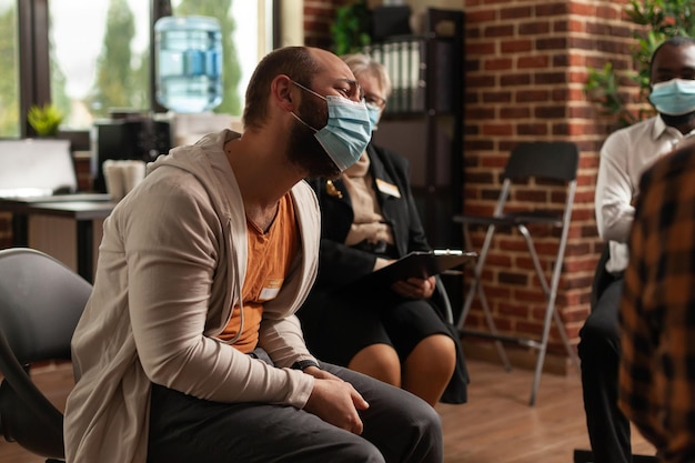 Free photo close up of man having conversation about addiction at aa group meeting with people during coronavorus pandemic. patients attending therapy session with psychologist, wearing face masks.