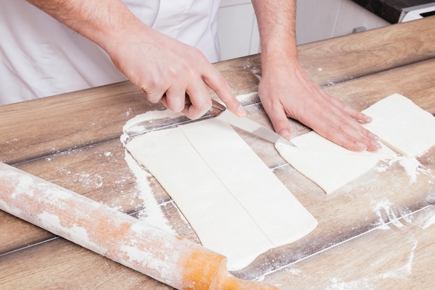 Close-up of man's hand cutting the rolled dough with knife on table
