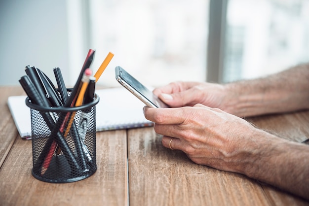 Foto gratuita il primo piano della mano dell'uomo che tiene lo smart phone a disposizione sopra lo scrittorio di legno