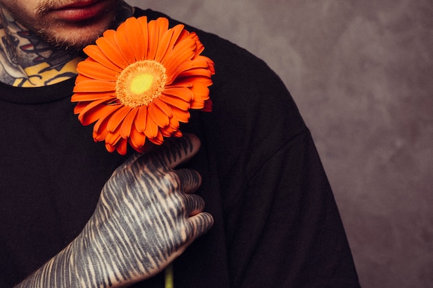 Close-up of a man with tattoo on his hand holding an orange gerbera flower over shoulder