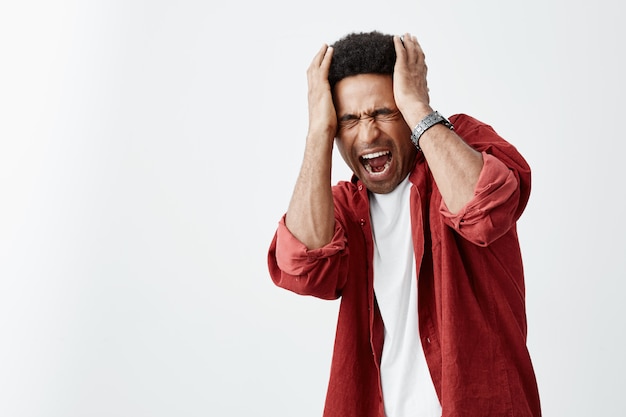 Close up of mature unhappy ta-skinned african man with curly hairstyle in white casual shirt and red shirt holding head with hands, struggling from headache after loud music at party.