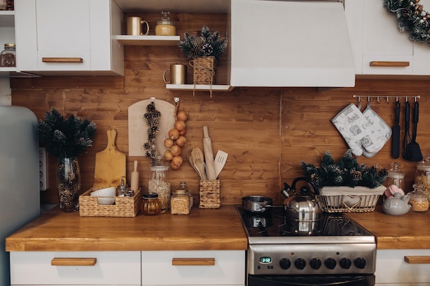 Крупный план современной уютной кухни в белых и коричневых тонах с вещами, плитой и декоративными еловыми ветками. Рождественские украшения.