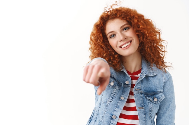 Close-up modern stylish ginger girl curly hair tilt head amused smiling broadly gentle kind grin pointing index finger camera make choice deciding you great candidate, standing white wall