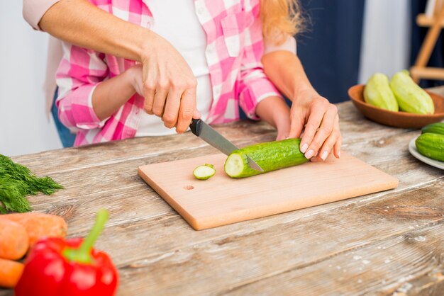 Close-up of mother holding the daughter's hand for cutting the cucumber on table